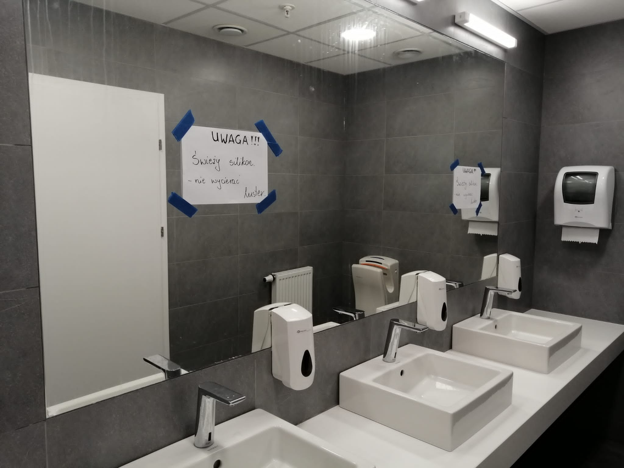 Publiczna toaleta z dwoma umywalkami, szarą glazurą i lustrem z kartką 'Uwaga! Świeży silikon - nie wycierać luster.'