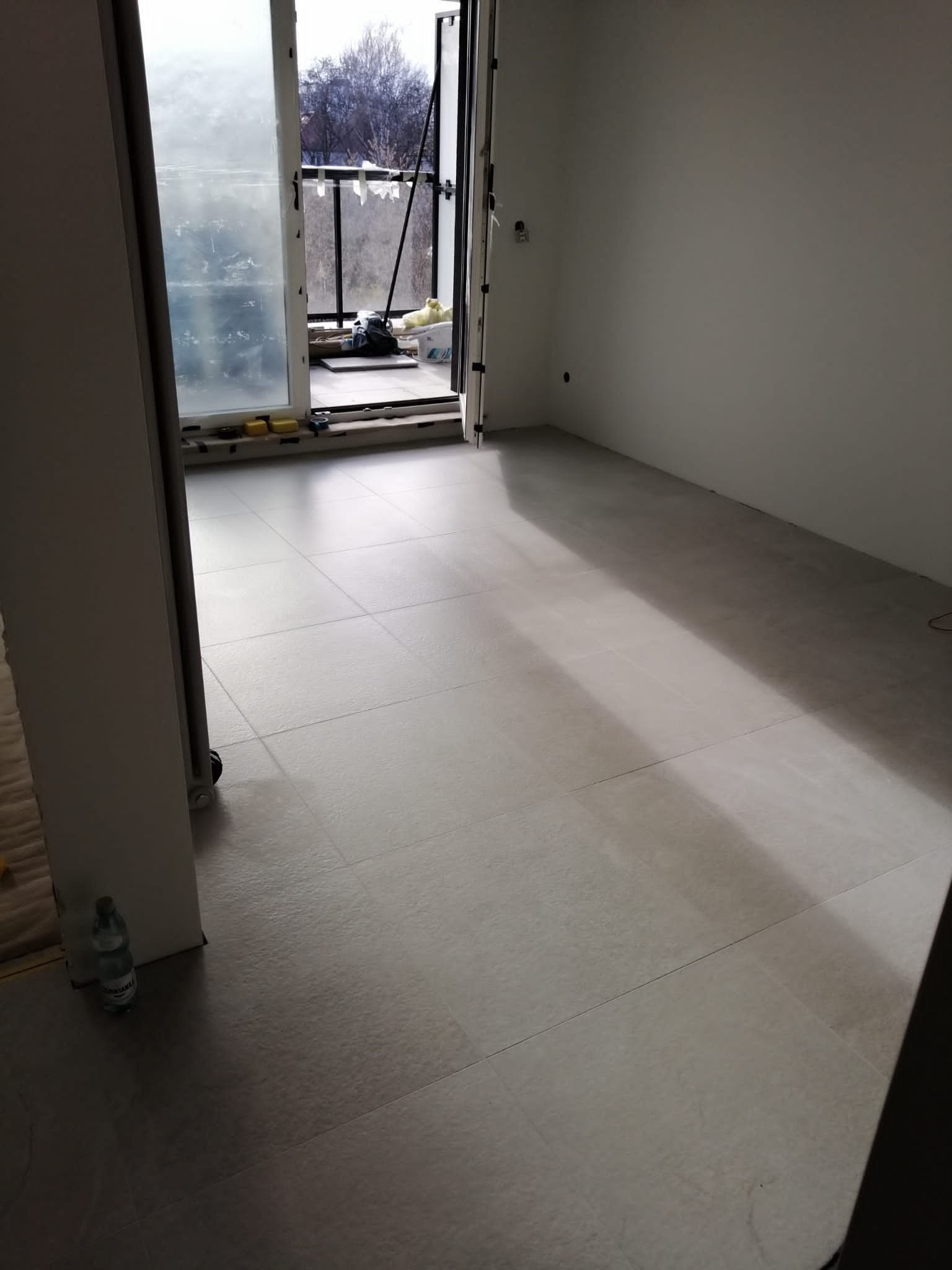 Ułożona podłoga z płytek w jasnoszarym odcieniu, widok z niskiego kąta, z otwartym balkonem w tle i butelką wody w rogu pomieszczenia.