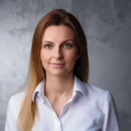 Portret kobiety z długimi, prostymi włosami w odcieniach brązu i blondu, ubranej w białą koszulę na tle szarej, teksturowanej ściany.