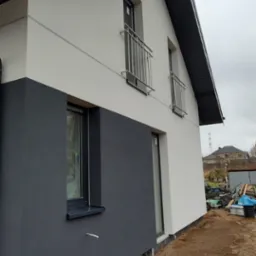 Dwukolorowa elewacja domu jednorodzinnego z balkonami, wykończona w odcieniach szarości i bieli, z widoczną warstwą izolacyjną przy gruncie.