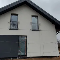 Dwukondygnacyjny dom z częściowo pomalowaną elewacją na szaro i biało, z czarnym dachem i stalowymi balustradami balkonów, zabezpieczony folią okienną, widoczne materiały budowlane.