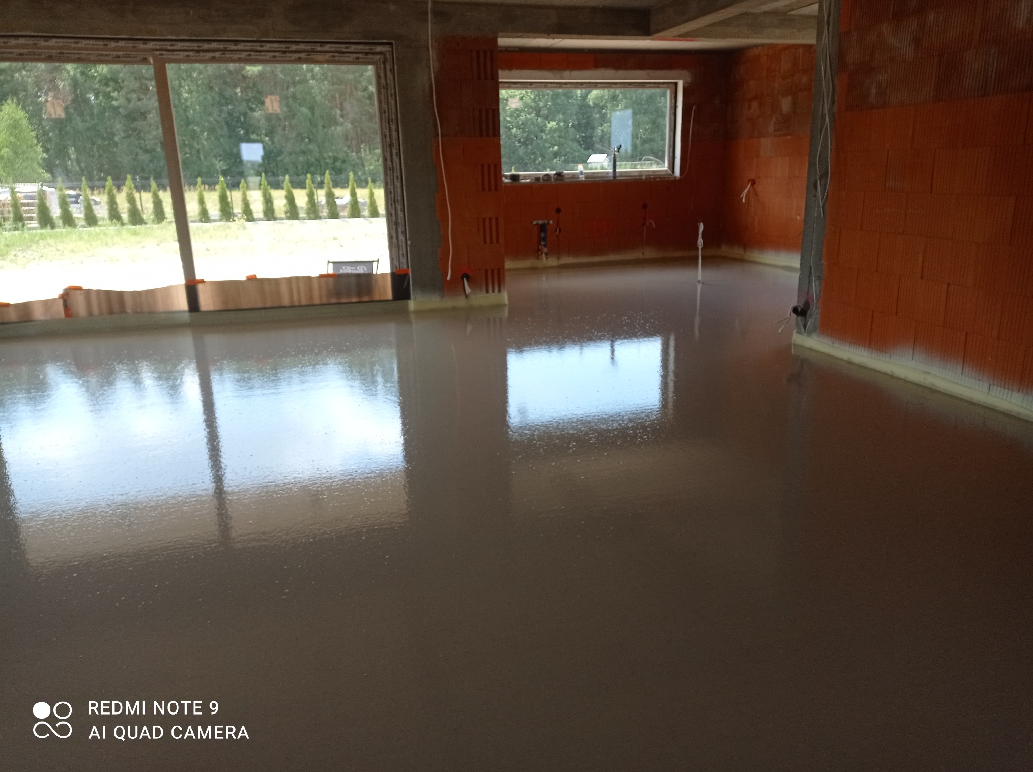 Świeżo wylana, gładka posadzka w surowym stanie, widoczne okna z widokiem na zieleń, ściany z czerwonej cegły dziurawki, przygotowane instalacje elektryczne.