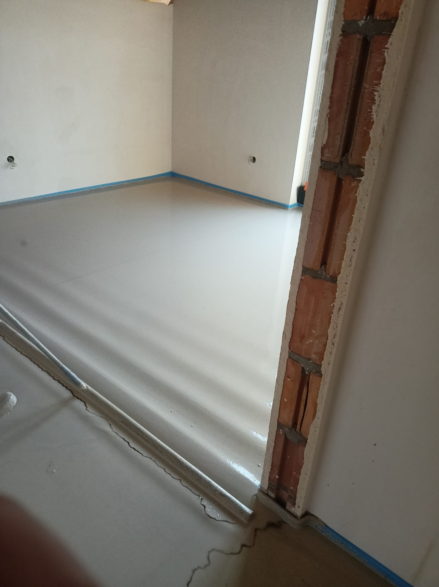 Świeżo wylana, gładka posadzka w jasnym pomieszczeniu zabezpieczona niebieską taśmą, widoczna rurka doprowadzająca materiał oraz fragment surowej ściany z cegieł.