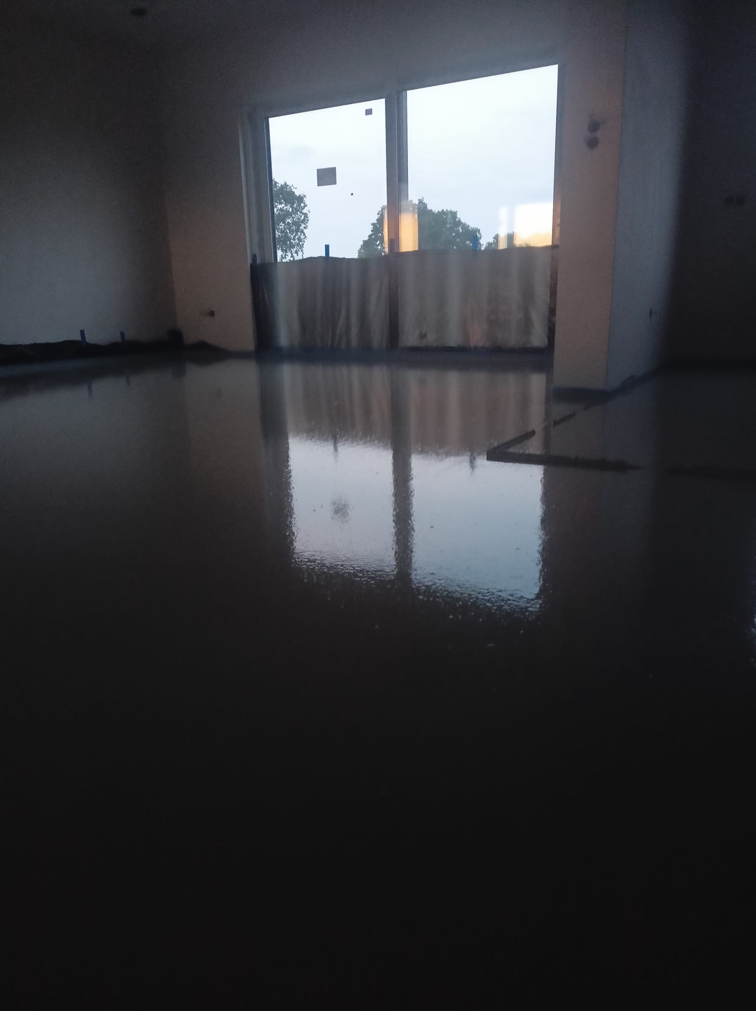 Świeżo wylana, błyszcząca posadzka w pomieszczeniu z dużym oknem, widoczne refleksy światła i niebieskie znaczniki poziomów na ścianach.