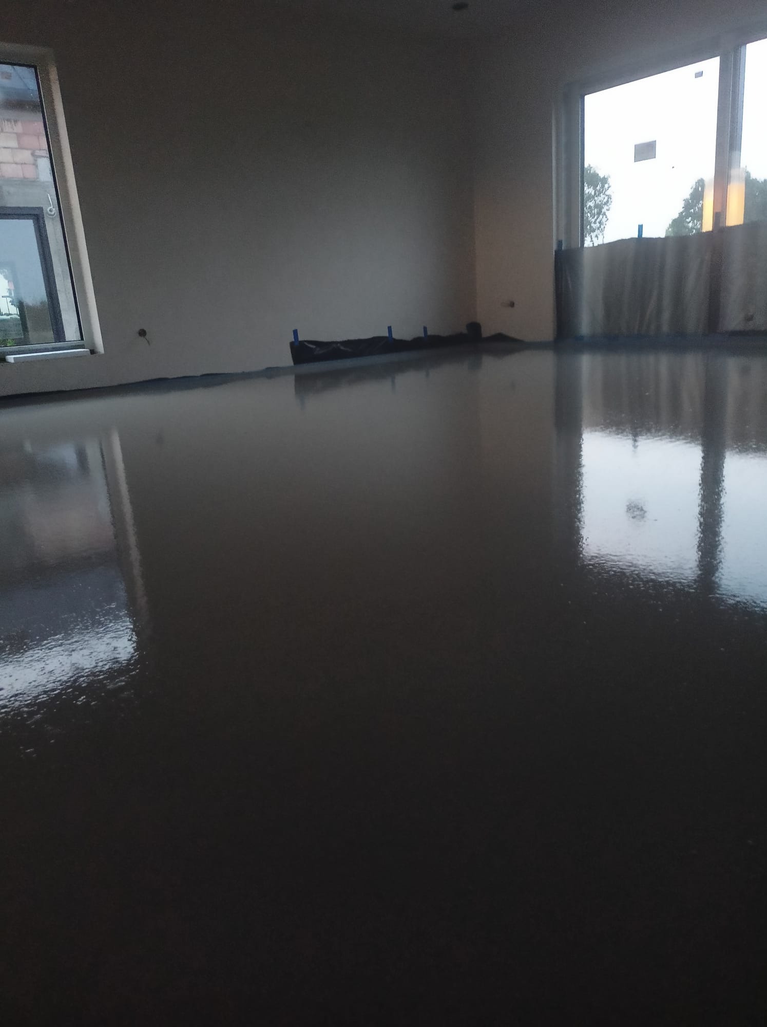 Świeżo wylana, gładka posadzka w jasnym pomieszczeniu z dużymi oknami, zabezpieczona folią przy ścianach.