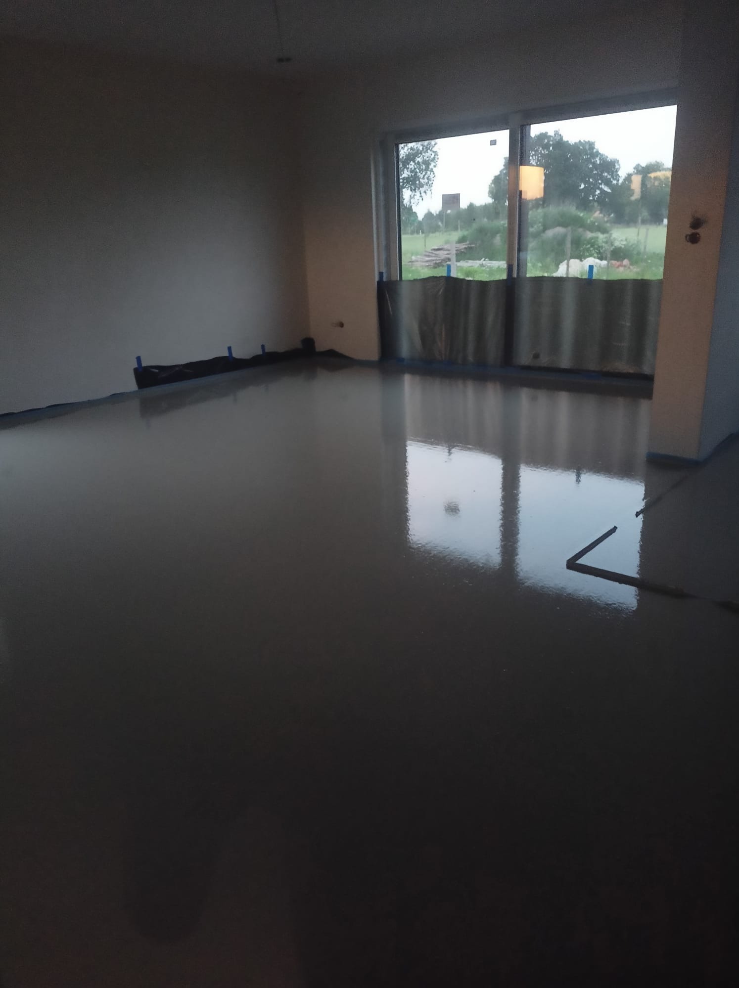 Świeżo wylana, gładka, szara posadzka w pustym pomieszczeniu z dużym oknem, zabezpieczonym folią malarską.