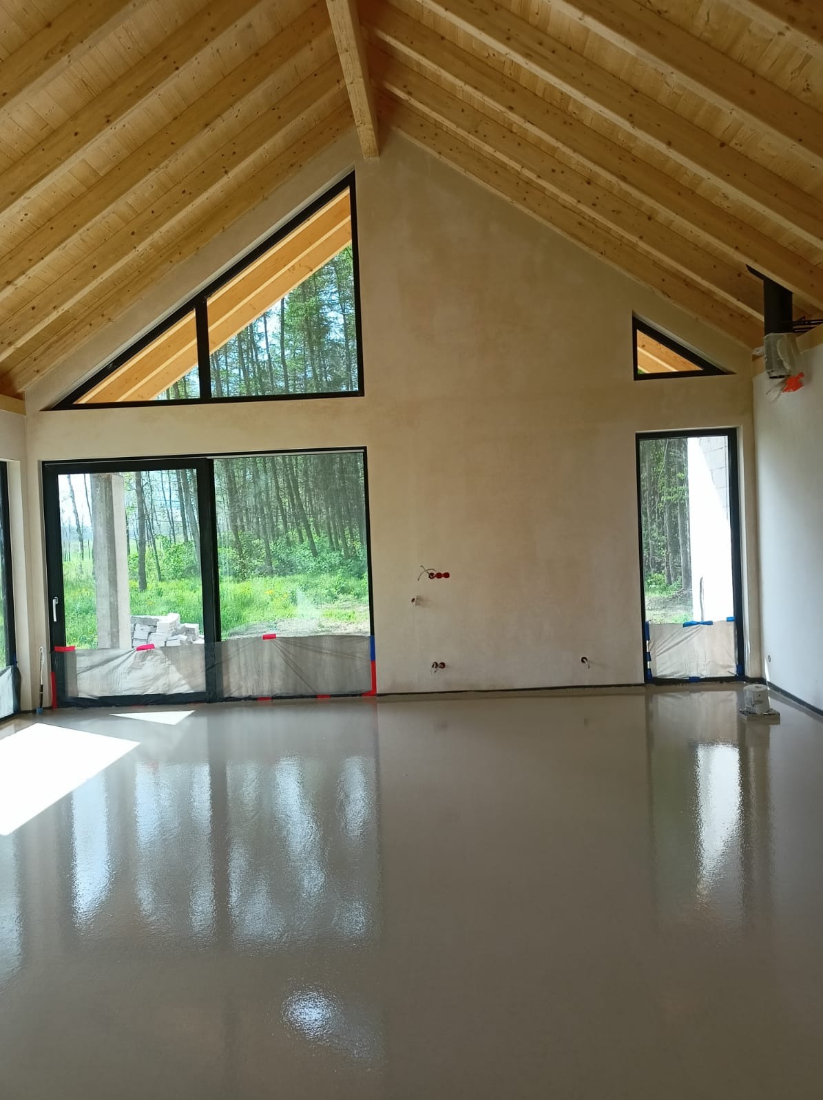 Świeżo wylana, gładka posadzka w jasnym pomieszczeniu z drewnianym stropem i dużymi oknami z widokiem na las. Posadzka zabezpieczona folią przy oknach i drzwiach.