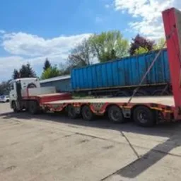 Ciężarówka niskopodwoziowa z niebieskim kontenerem na platformie, zaparkowana na betonowym placu w słoneczny dzień, widoczne drzewa w tle.