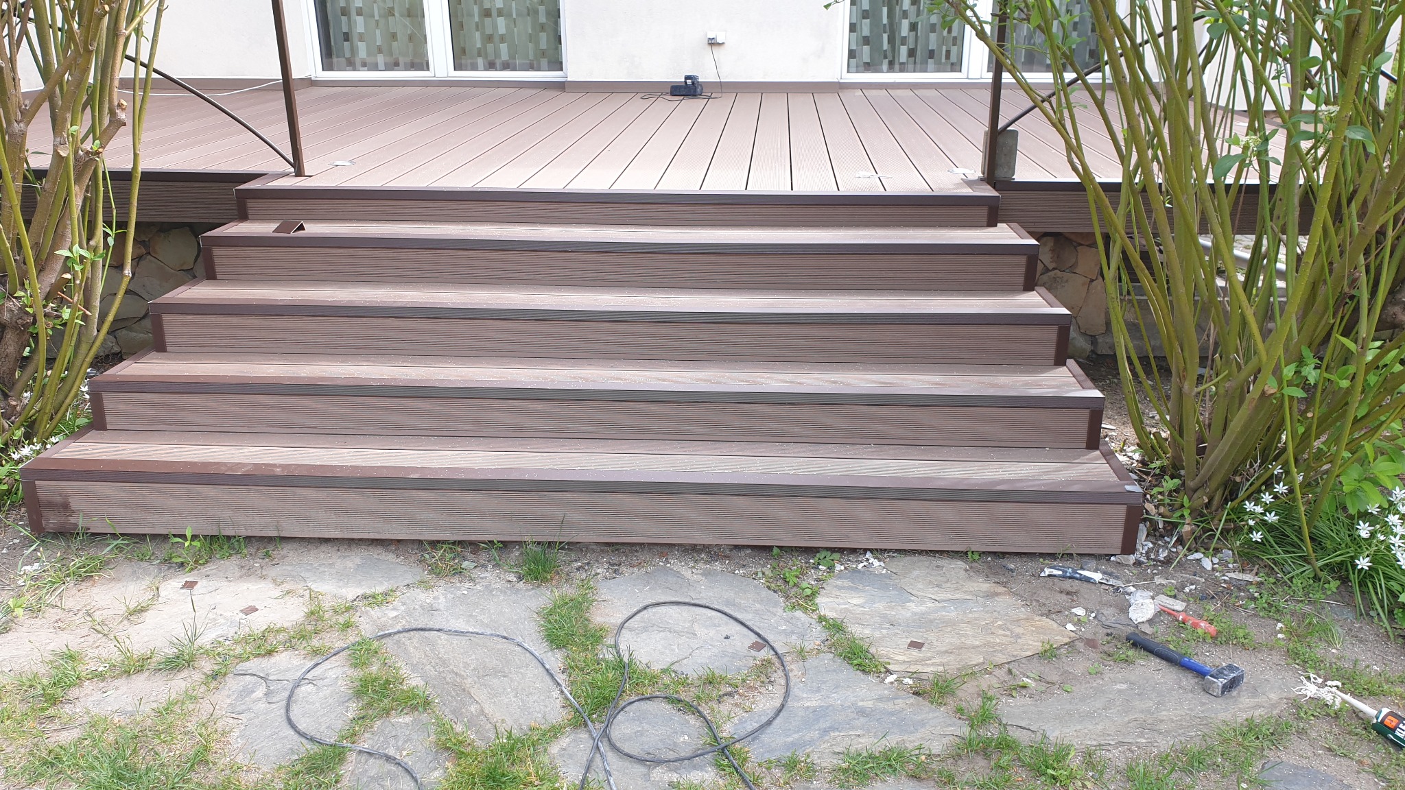 Schody z deski kompozytowej prowadzące na taras, widoczne narzędzia montażowe i fragment roślinności.