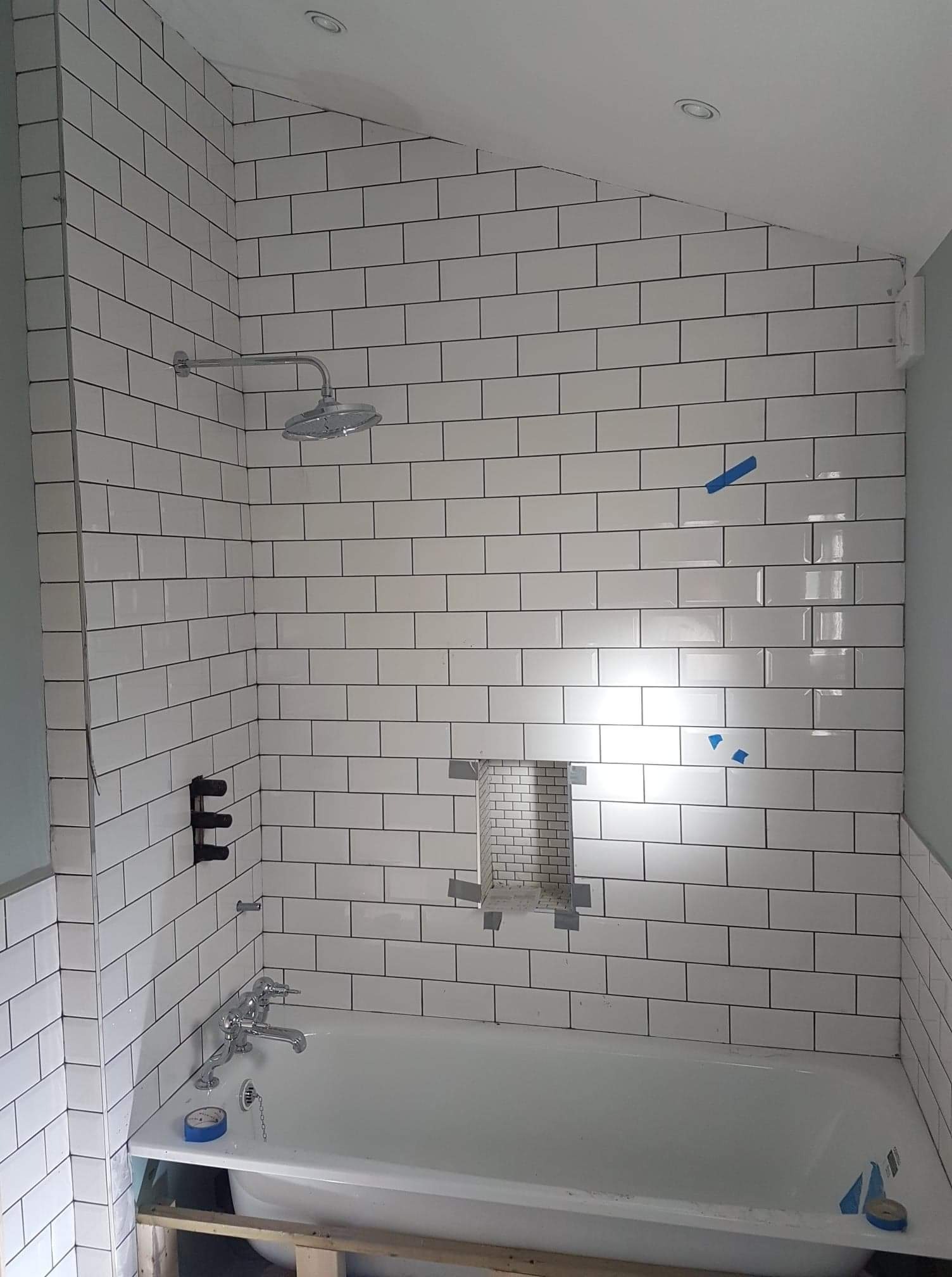 Białe, prostokątne płytki z czarną fugą w łazience podczas montażu, widoczna wanna, bateria, prysznic i wnęka wyłożona drobną mozaiką, zabezpieczona taśmą malarską.