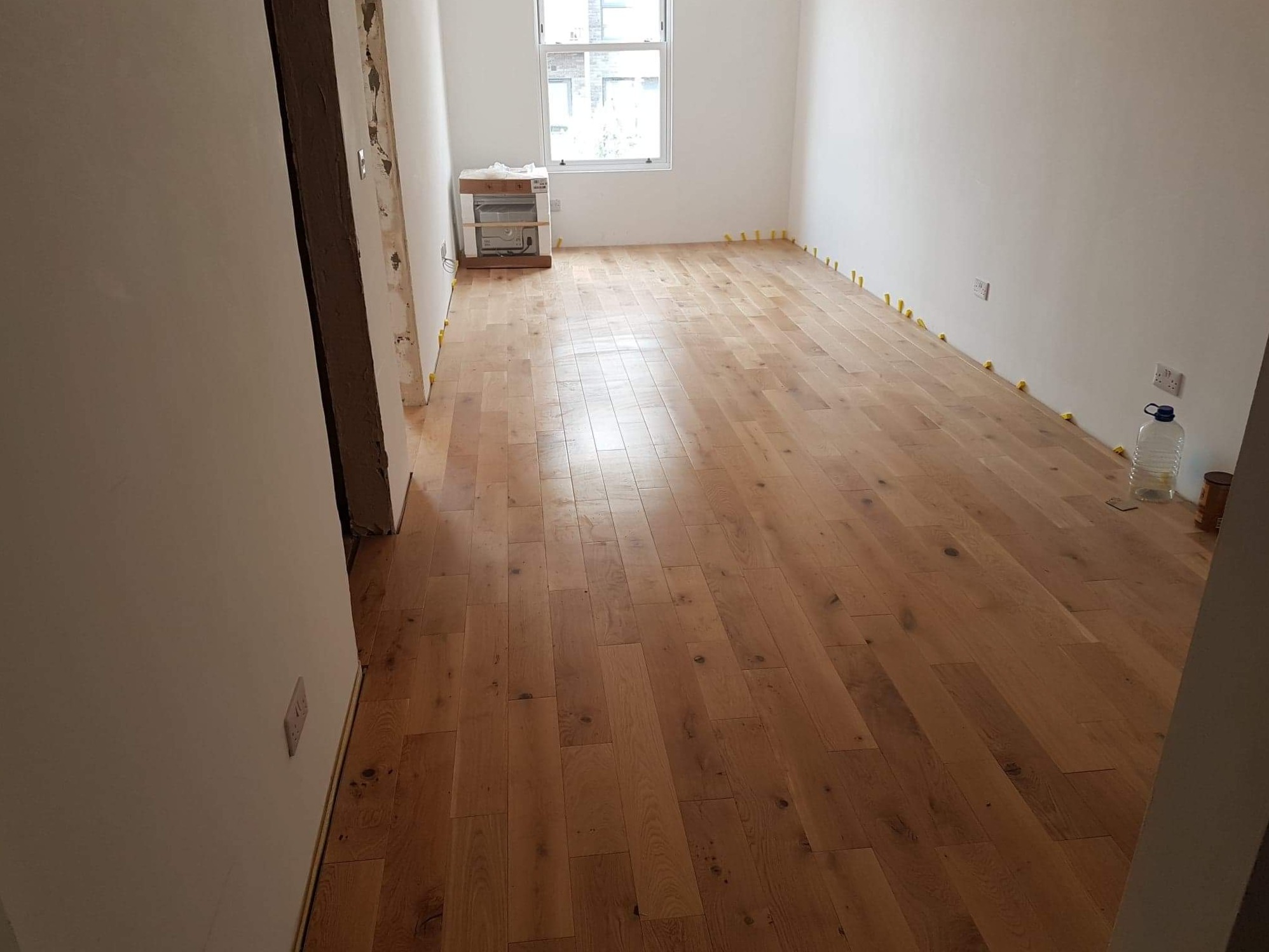 Pokój po remoncie z nową drewnianą podłogą, zabezpieczoną żółtymi klinami dystansowymi przy ścianach, widoczne gniazdka elektryczne i okno z widokiem na budynki.