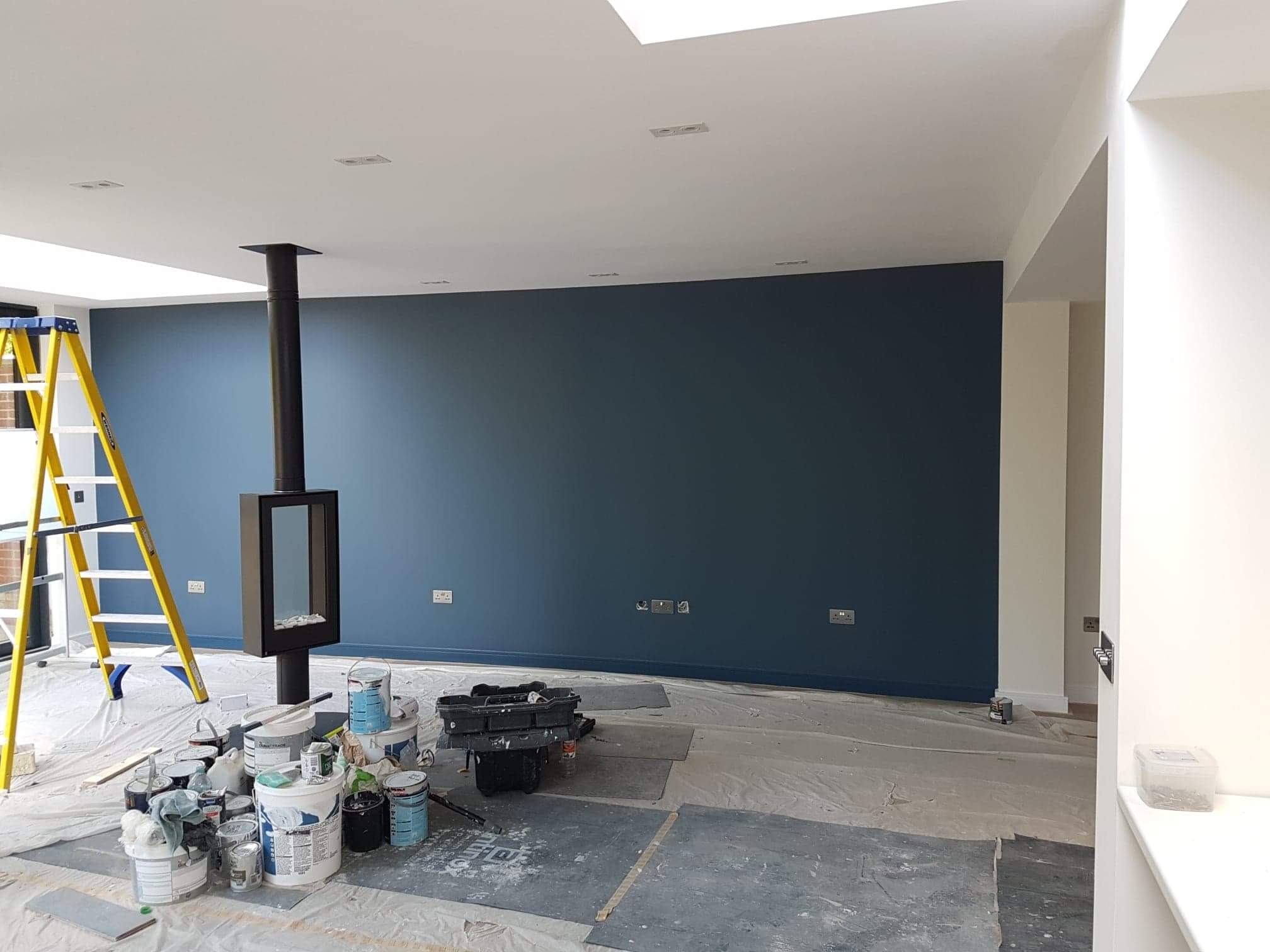 Świeżo pomalowana ściana w odcieniu głębokiego granatu w przestronnym salonie z nowoczesnym kominkiem, zabezpieczona folią podłoga i żółta drabina malarska, widoczne puszki z farbą i narzędzia...