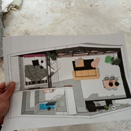 Andrii Mantelchuk - Rzut z góry na plan mieszkania z zaznaczonym układem mebli i wyposażenia, trzymany w dłoni na tle surowego wykończenia wnętrza.