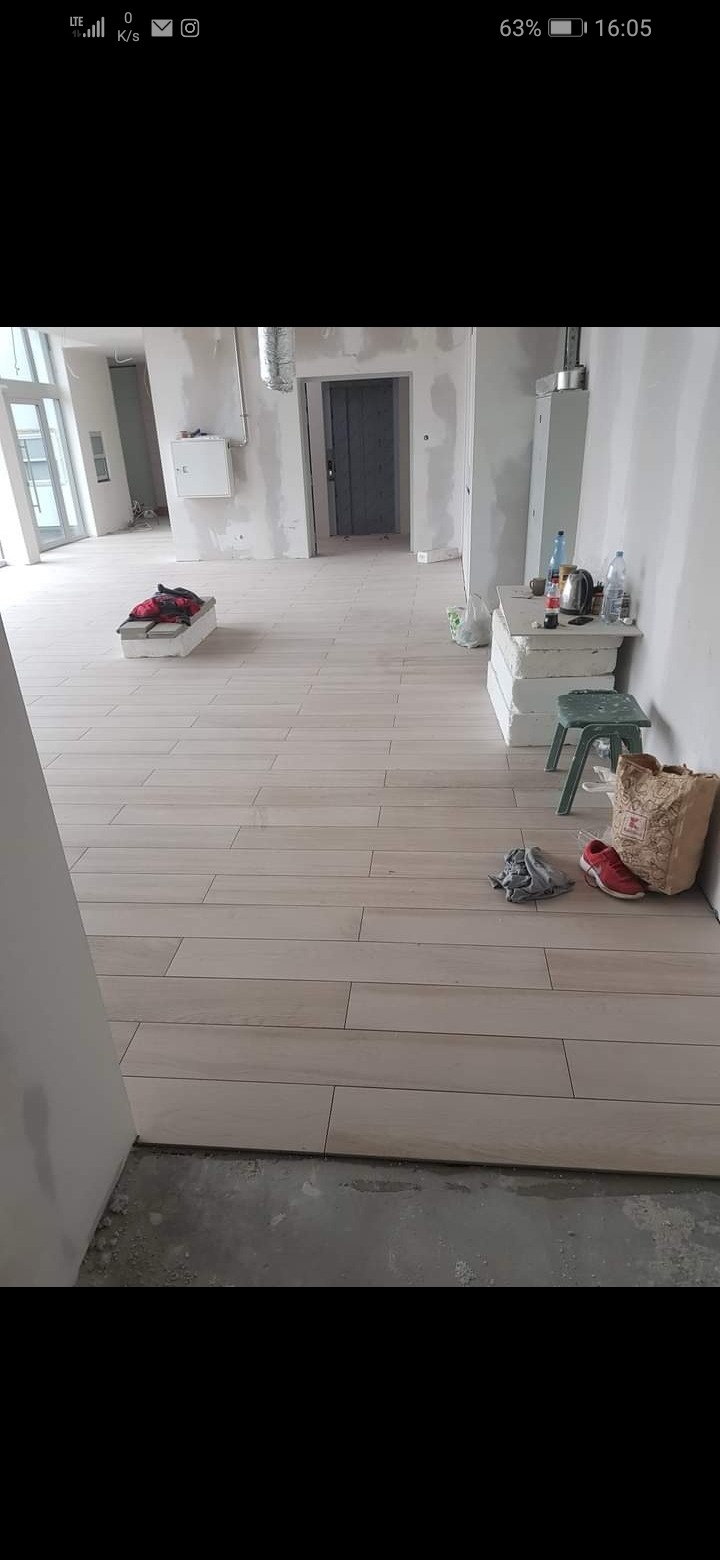 Ułożona podłoga z płytek imitujących drewno w surowym wnętrzu, widoczne przygotowania do dalszych prac wykończeniowych, materiały budowlane i osobiste rzeczy pracowników.