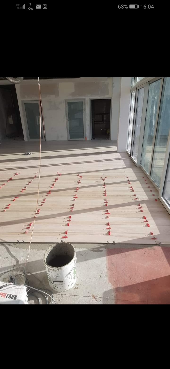 Układanie jasnych paneli podłogowych z użyciem czerwonych klipsów poziomujących w jasnym, przestronnym wnętrzu z dużymi oknami i surowymi ścianami.