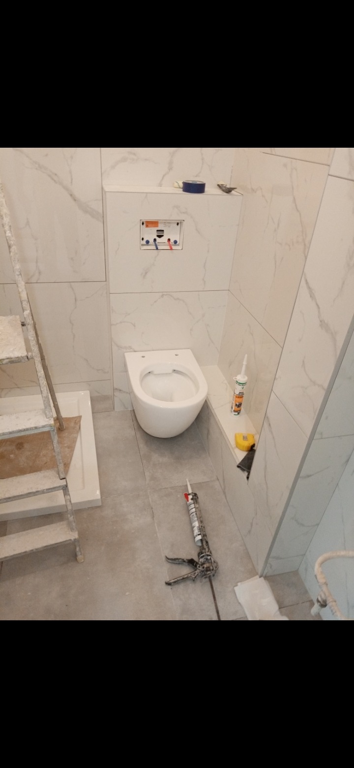 Wnętrze łazienki w trakcie wykańczania: białe marmurowe płytki ścienne, podwieszana toaleta bez deski, drabina malarska, pistolet do silikonu, tuba silikonu i poziomica.