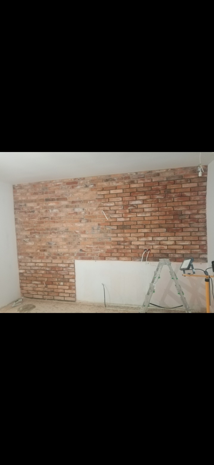Ściana z cegły dekoracyjnej w trakcie montażu, widoczne niedokończone fragmenty i drabina z oświetleniem roboczym.