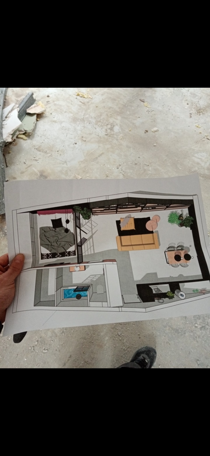 Rzut z góry na plan mieszkania z zaznaczonym układem mebli i wyposażenia, trzymany w dłoni na tle surowego wykończenia wnętrza.