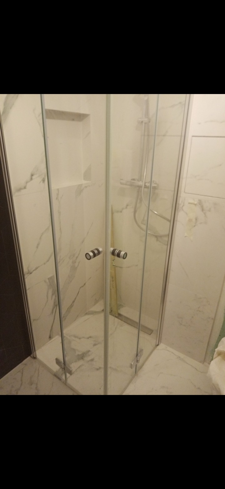 Kabina prysznicowa narożna z przezroczystego szkła, z chromowanymi uchwytami, zamontowana w łazience wykończonej płytkami imitującymi marmur, widoczny odpływ liniowy.