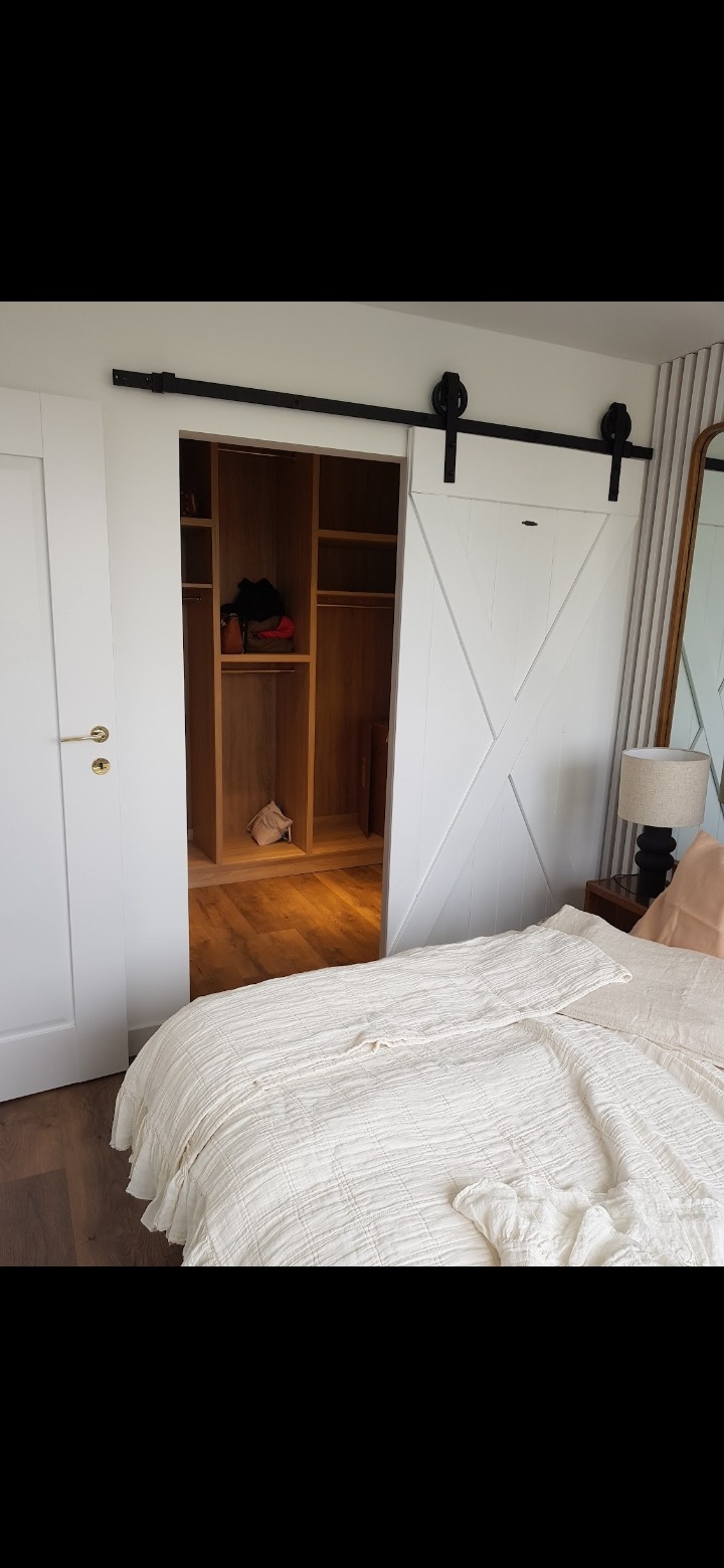 Białe, przesuwne drzwi stodołowe z czarnym okuciem, prowadzące do drewnianej garderoby w sypialni z łóżkiem w stylu skandynawskim.