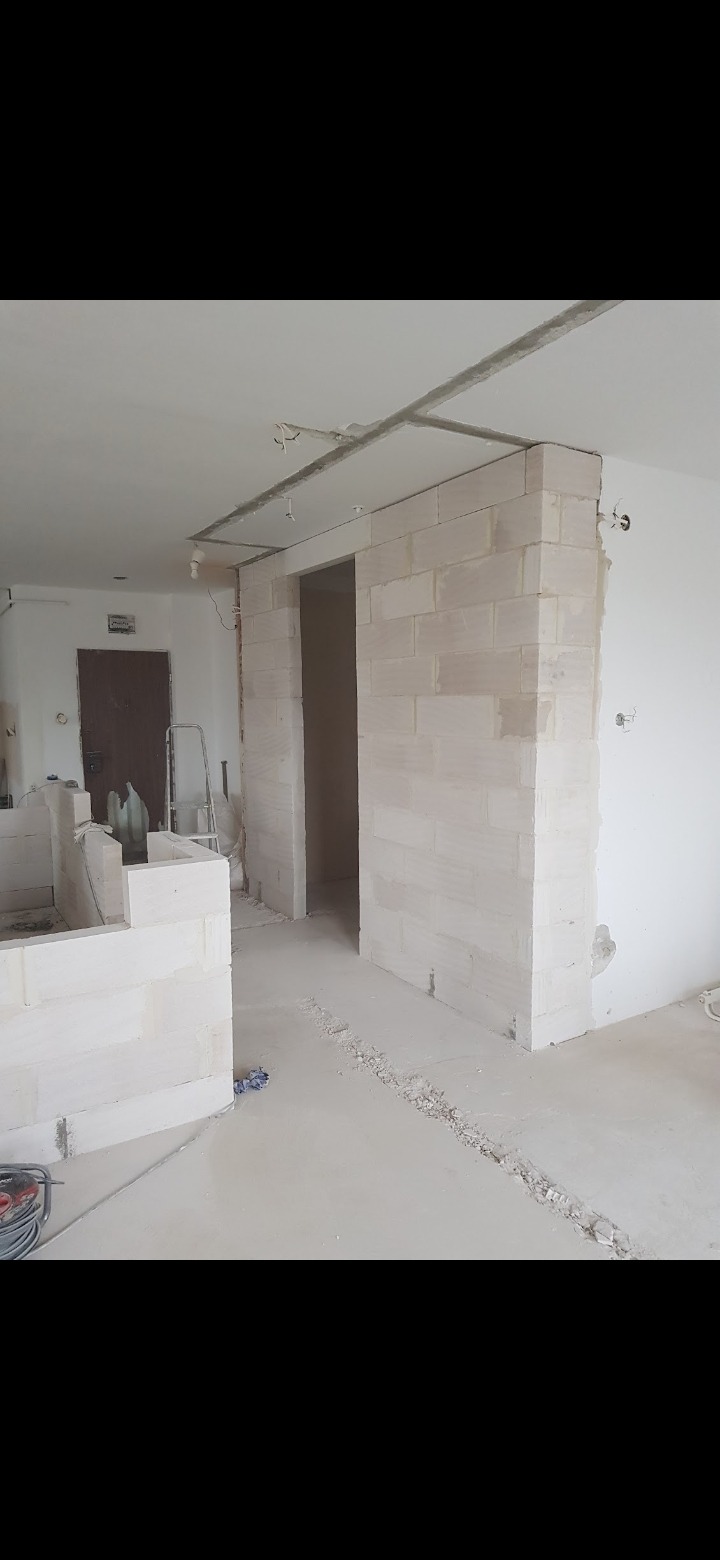 Wnętrze w trakcie prac murarskich z bloków, widoczne fragmenty ścian i otwory drzwiowe, surowy beton na podłodze, instalacja elektryczna na suficie, drabina oparta o ścianę w tle.