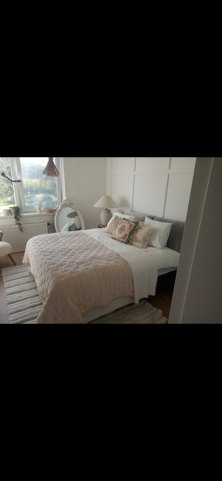 Jasna sypialnia z łóżkiem przykrytym pudrowo-różową narzutą, dekoracyjnymi poduszkami, owalnym lustrem w ozdobnej ramie i widokiem na ogród za oknem.