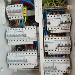 Otwarta skrzynka elektryczna z widocznymi bezpiecznikami, przewodami w różnych kolorach (niebieski, czerwony, żółty, zielony, czarny) i elementami mocującymi, ukazująca skomplikowaną instalację...