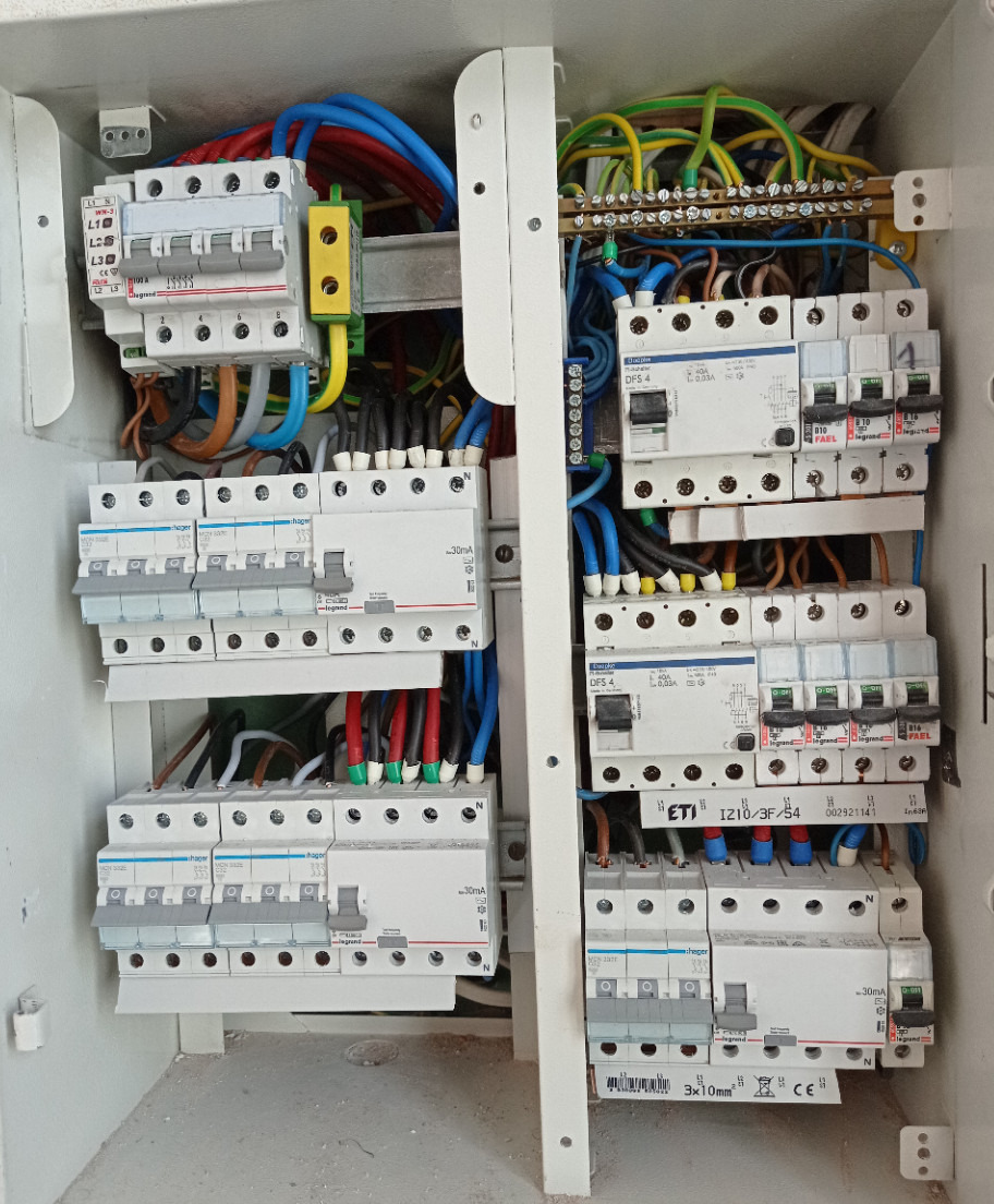 Otwarta skrzynka elektryczna z widocznymi bezpiecznikami, przewodami w różnych kolorach (niebieski, czerwony, żółty, zielony, czarny) i elementami mocującymi, ukazująca skomplikowaną instalację...