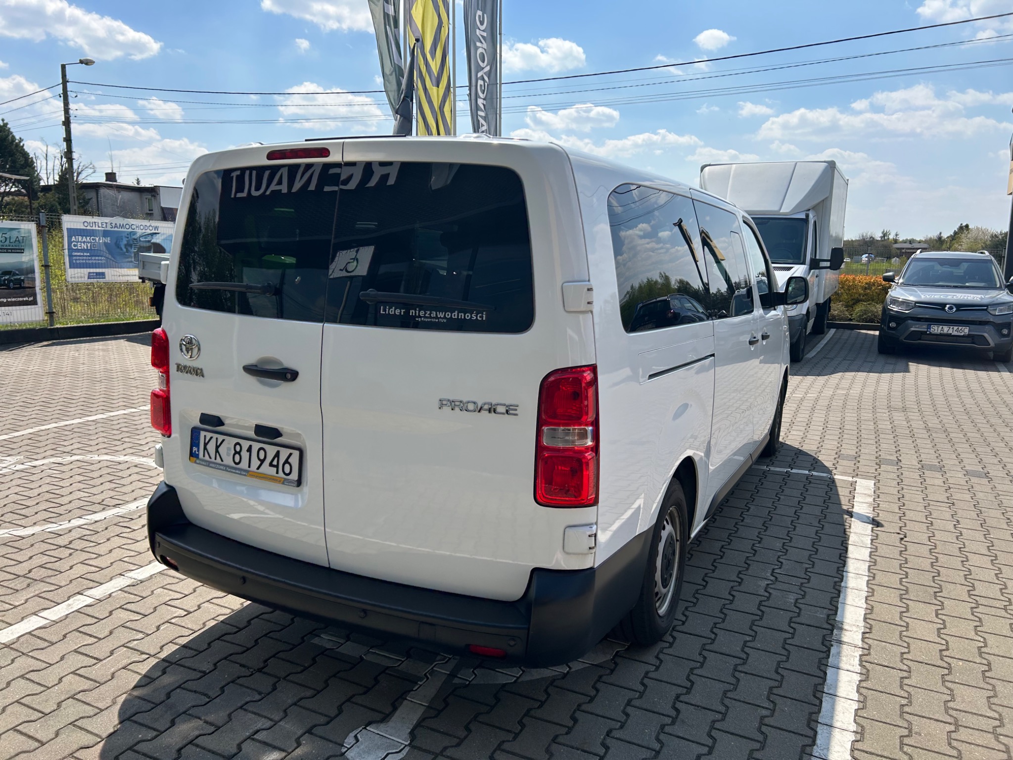 Biały samochód dostawczy marki Toyota Proace z widoczną naklejką 'Lider Niezawodności' na tylnej szybie, zaparkowany na parkingu z kostki brukowej w słoneczny dzień.