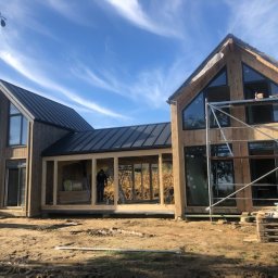 Good Wood House - Surowy, nowoczesny dom w trakcie budowy, z widoczną konstrukcją drewnianą, czarnym dachem i rusztowaniami, na tle błękitnego nieba z chmurami.