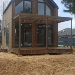 Nowoczesny, dwukondygnacyjny dom z ciemnego drewna z dużymi, trójkątnymi oknami na poddaszu i zadaszonym tarasem, widoczny komin ze stali nierdzewnej, na pierwszym planie piaszczysty teren.