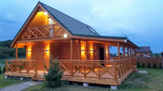 Nowoczesny dom z drewna, z podświetlonym balkonem i werandą, otoczony zielenią, sfotografowany o zmierzchu.