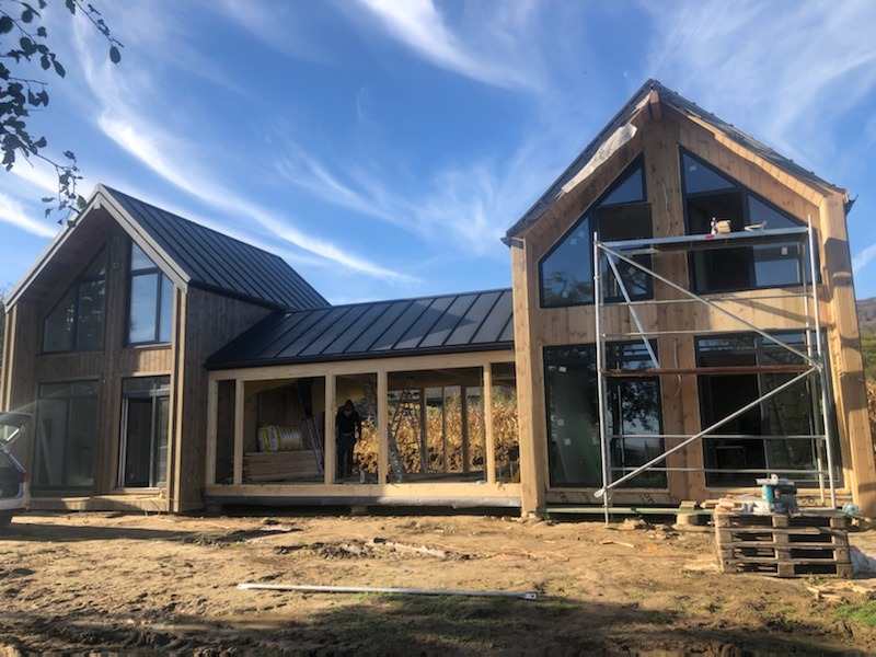 Surowy, nowoczesny dom w trakcie budowy, z widoczną konstrukcją drewnianą, czarnym dachem i rusztowaniami, na tle błękitnego nieba z chmurami.