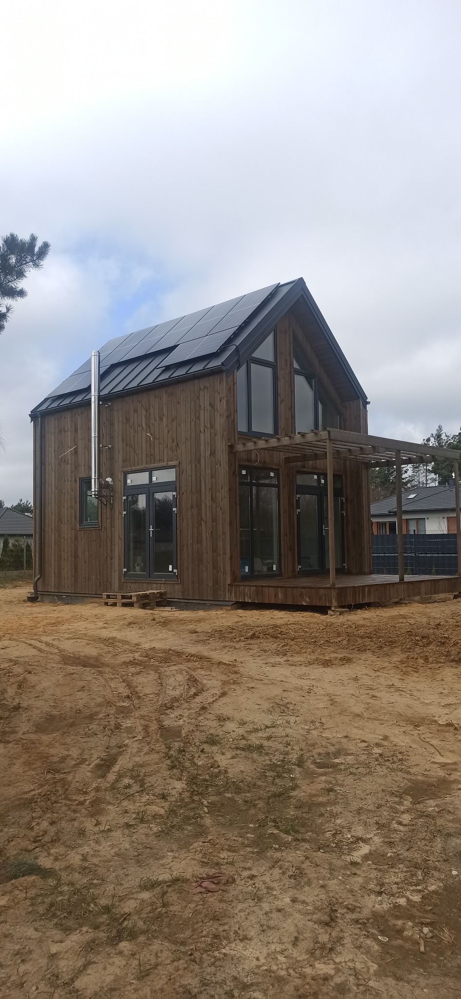 Nowoczesny dom z ciemnego drewna z panelami słonecznymi na dachu i drewnianym tarasem, widoczny komin ze stali nierdzewnej i paleta pod oknem, otoczenie w stanie surowym.