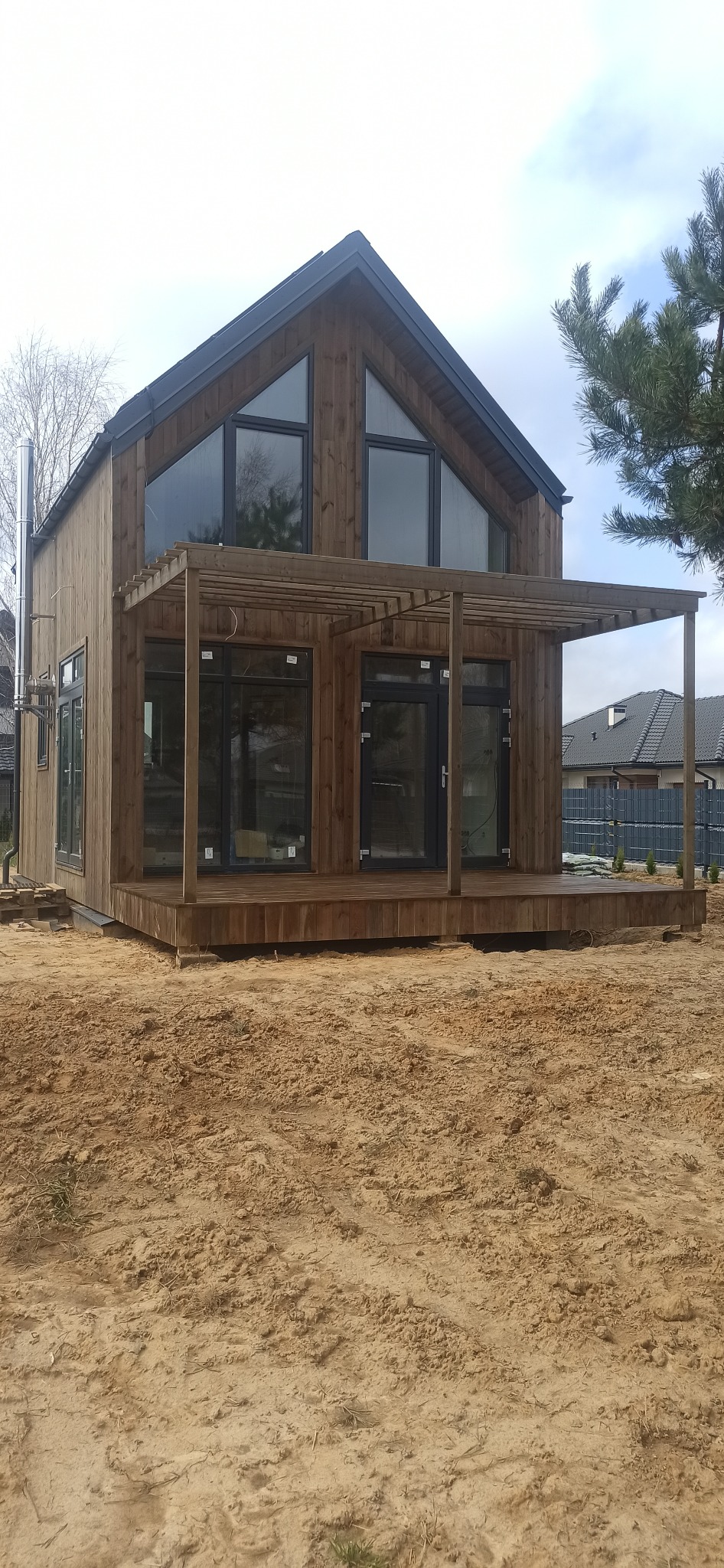 Nowoczesny, dwukondygnacyjny dom z ciemnego drewna z dużymi, trójkątnymi oknami na poddaszu i zadaszonym tarasem, widoczny komin ze stali nierdzewnej, na pierwszym planie piaszczysty teren.