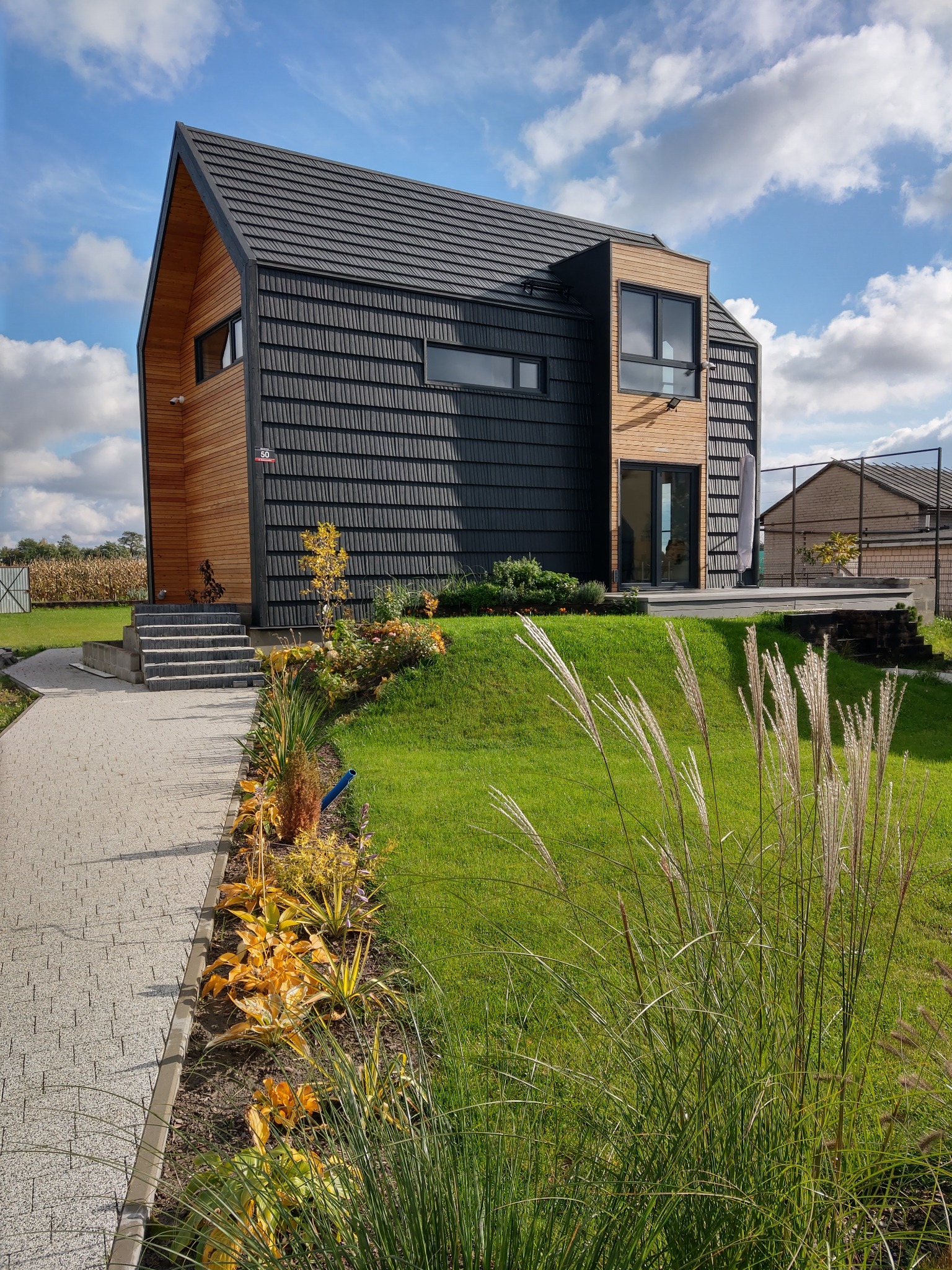 Nowoczesny dom z drewnianą elewacją i ciemną dachówką, otoczony zielonym trawnikiem i ozdobnymi roślinami, widok z poziomu ścieżki prowadzącej do wejścia.