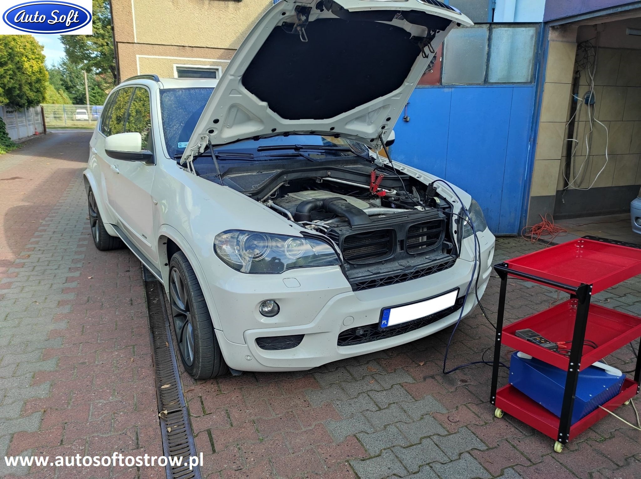 Biały SUV z otwartą maską, podłączony do akumulatora klemami, obok wózek warsztatowy z urządzeniem diagnostycznym, widoczne elementy silnika.