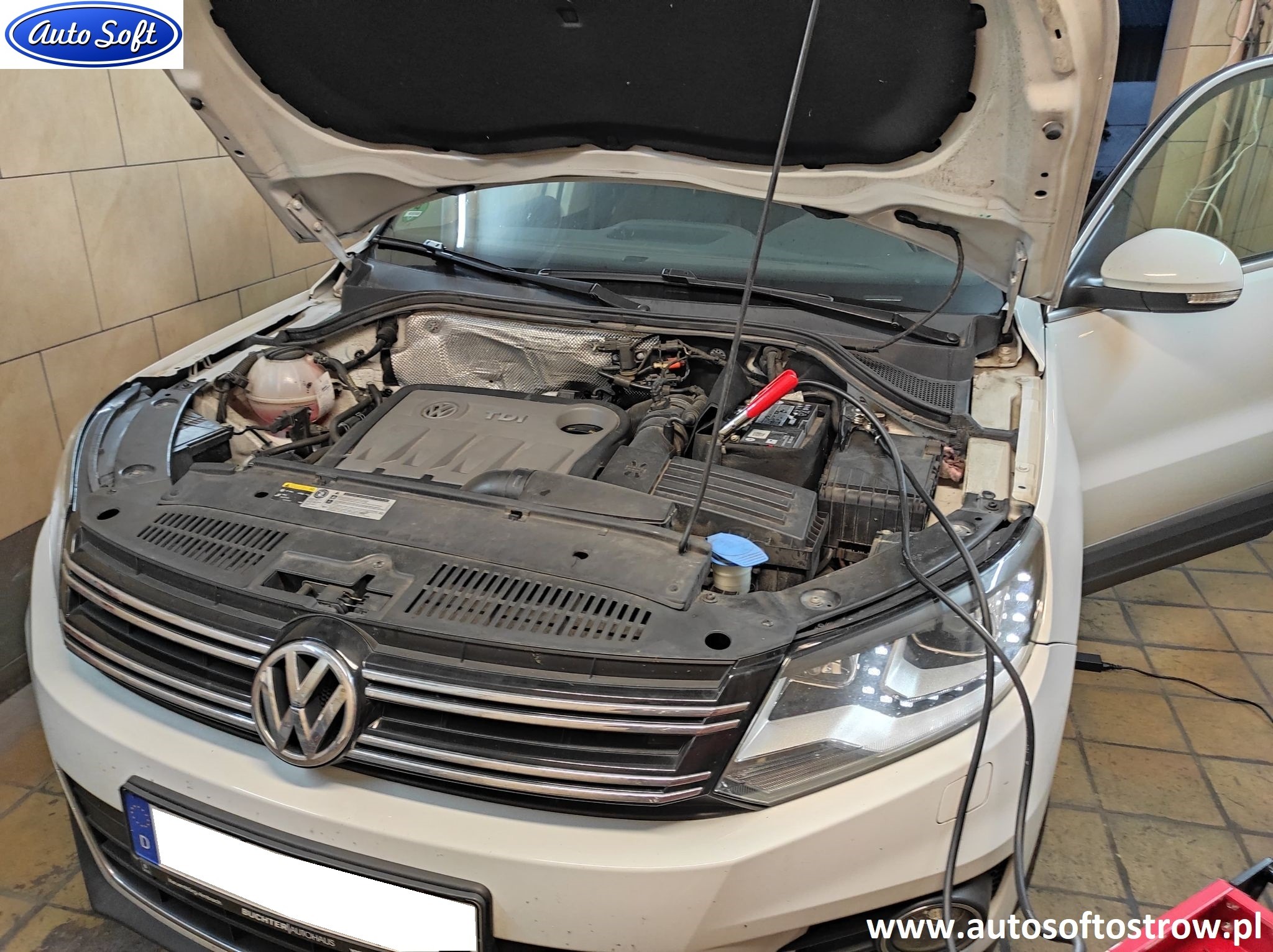Otwarta maska białego samochodu Volkswagen Tiguan, widoczny silnik z podłączonymi przewodami diagnostycznymi i klemami akumulatora. Na masce widoczne logo firmy Auto Soft.