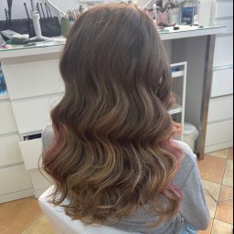 Malapert Beauty Studio - Falowane, brązowe włosy z różowymi pasemkami na końcach, widok z tyłu, w tle stanowisko z akcesoriami.
