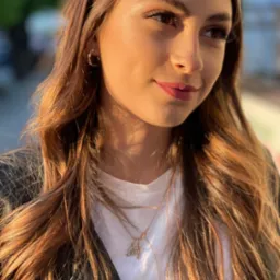 Portret młodej kobiety z lekkim, naturalnym makijażem, długimi, brązowymi włosami układającymi się w fale, oświetlonymi ciepłym, bocznym światłem słonecznym, z widocznym delikatnym błyskiem...