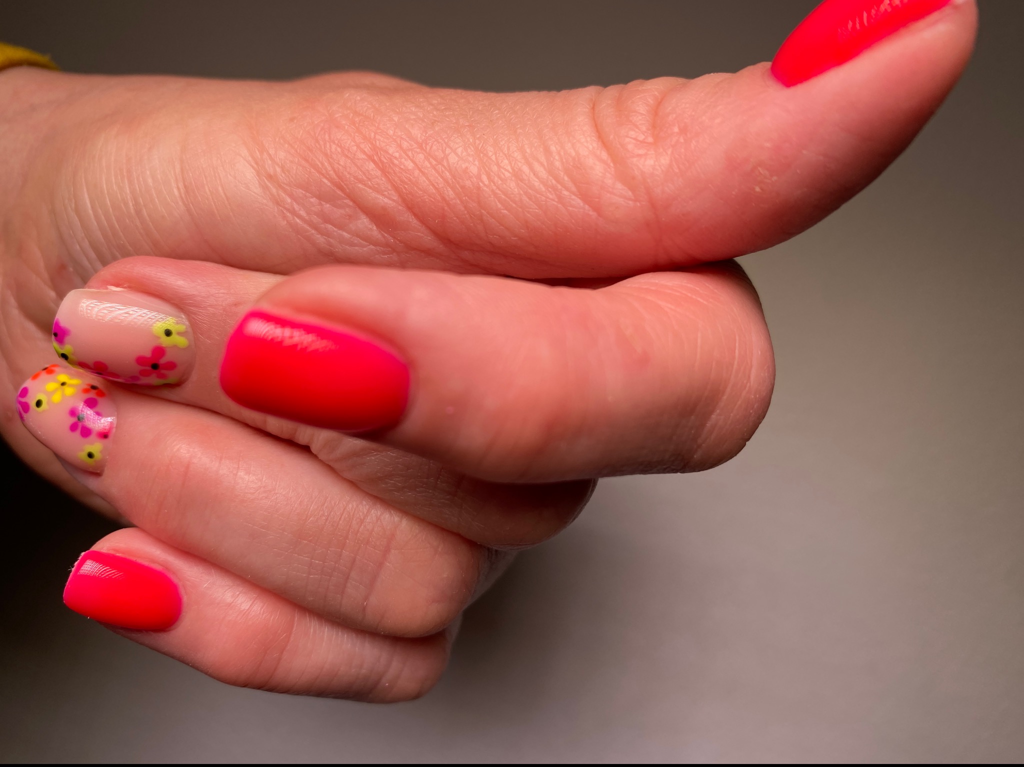 Dłoń z pomalowanymi paznokciami: trzy czerwone, jeden beżowy z drobnymi kwiatkami w kolorach różu, żółci i czerni.