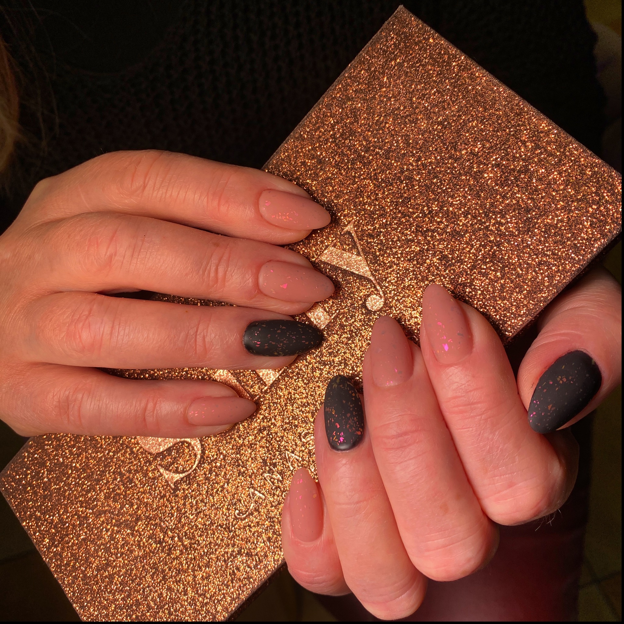 Dłonie prezentują paznokcie: trzy w odcieniu nude z różowym brokatem, dwa matowe czarne z kolorowymi drobinkami, na tle błyszczącej, złotej palety do cieni.