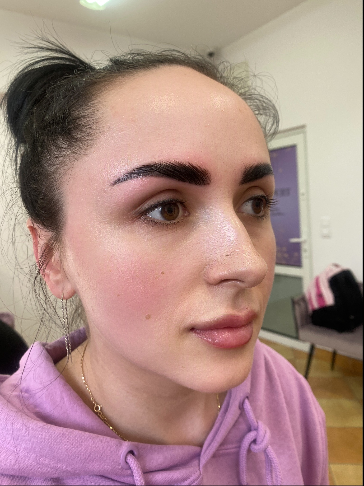 Profil kobiety po zabiegu stylizacji brwi, z widocznymi świeżo wyregulowanymi, ciemnymi brwiami i delikatnym makijażem ust, w tle niewyraźne elementy wyposażenia salonu.