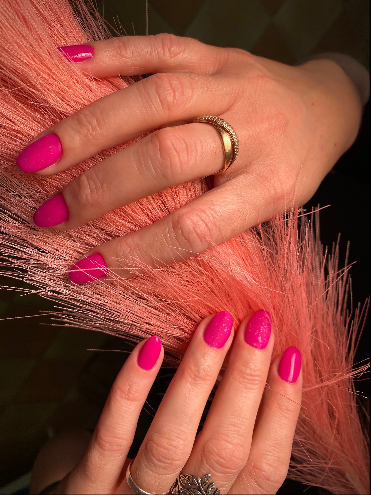 Dłonie z intensywnie różowym manicure hybrydowym, ozdobione złotym pierścionkiem, trzymające dekoracyjną, koralową trawę.