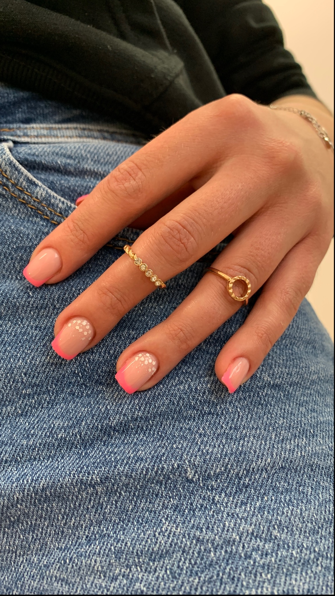 Dłoń z różowym frenczem i białymi kropkami na dwóch paznokciach, ozdobiona dwoma złotymi pierścionkami na tle dżinsu.