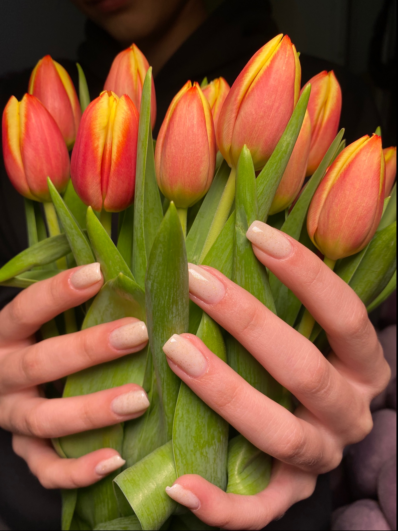 Dłonie z beżowym manicure z drobnymi złotymi drobinkami trzymają bukiet czerwono-żółtych tulipanów na ciemnym tle.