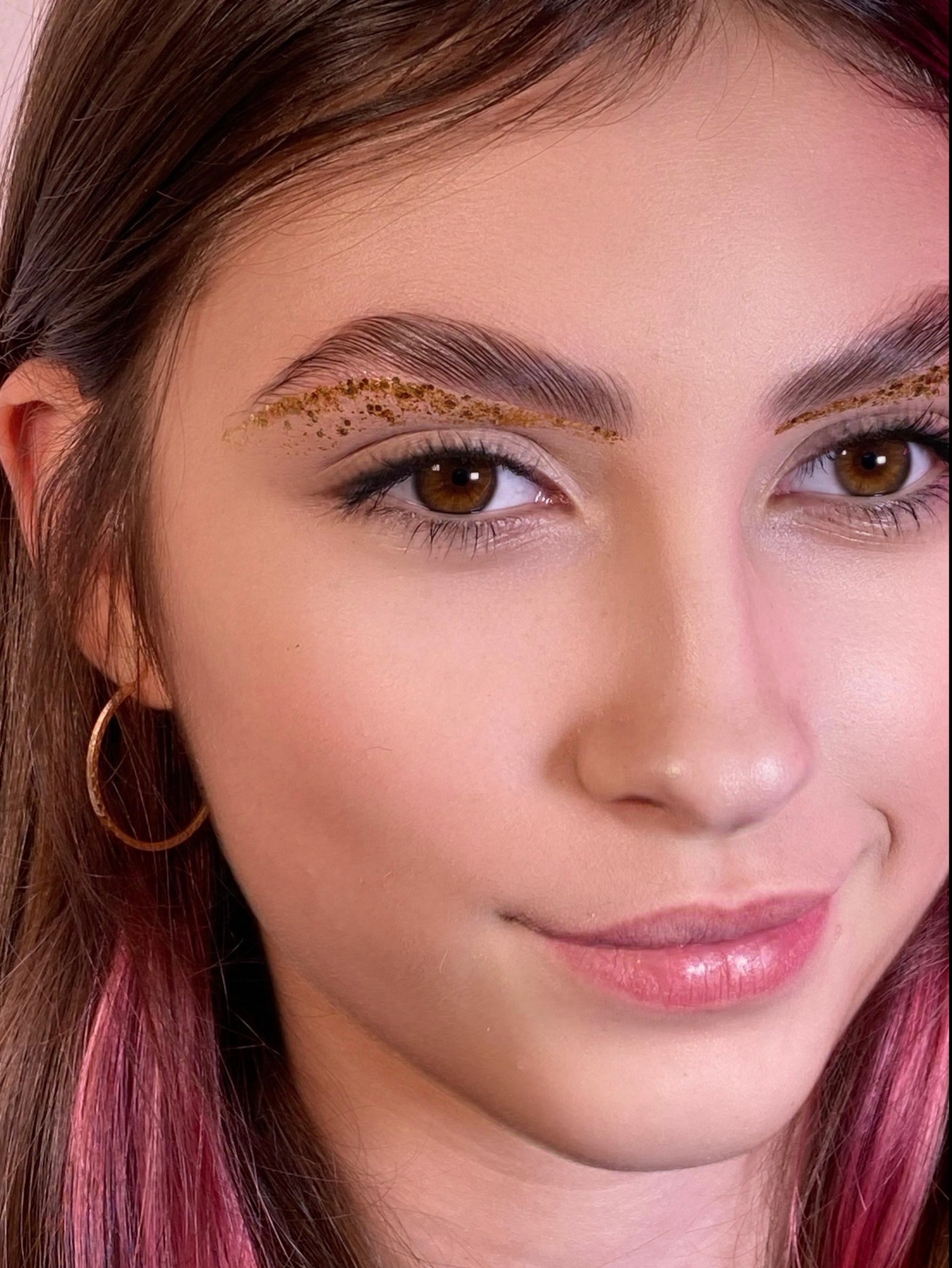 Portret zbliżeniowy młodej kobiety z brązowymi oczami i brwiami ozdobionymi złotym brokatem, różowe pasemka we włosach i błyszczące usta.