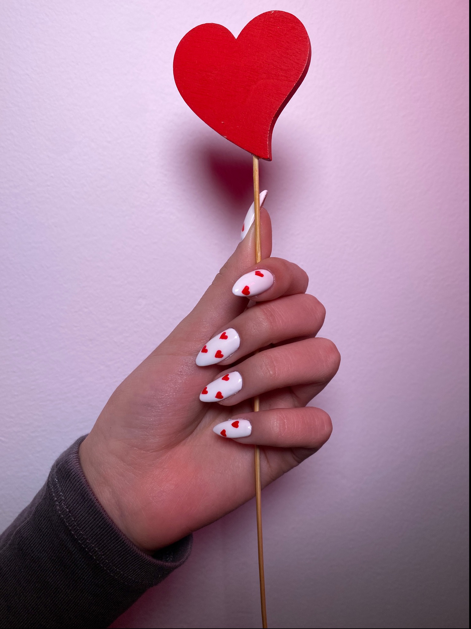Kobieca dłoń trzymająca drewniane czerwone serce na patyku, prezentująca białe paznokcie w kształcie migdała z drobnymi czerwonymi serduszkami na tle jasnoróżowej ściany.