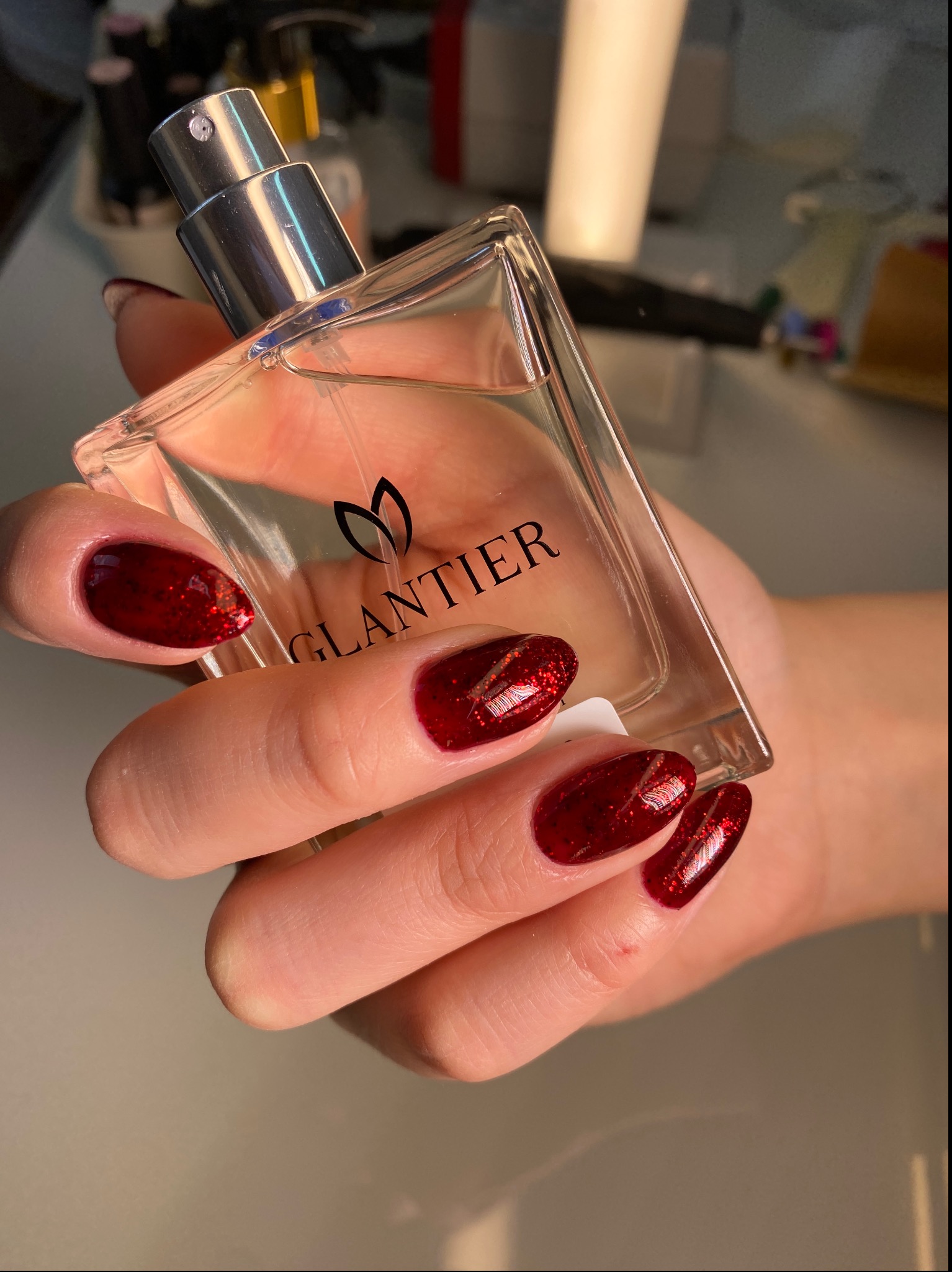 Dłoń z czerwonym, brokatowym manicure hybrydowym trzymająca flakon perfum Glantier o przezroczystej butelce i srebrnym atomizerze.