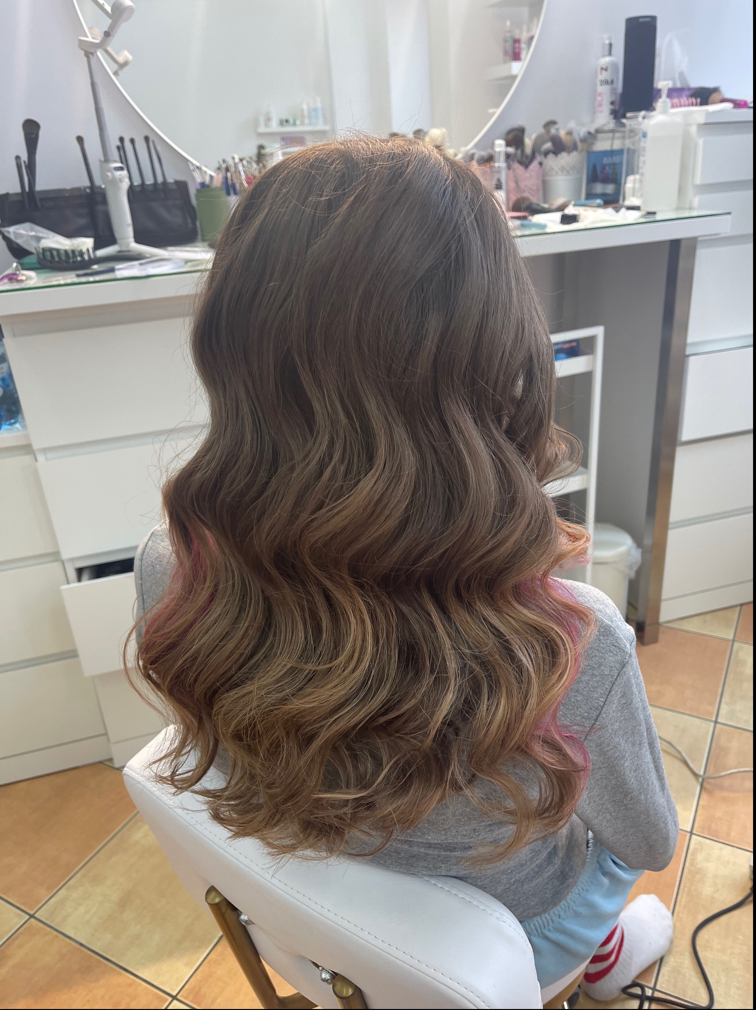 Falowane, brązowe włosy z różowymi pasemkami na końcach, widok z tyłu, w tle stanowisko z akcesoriami.